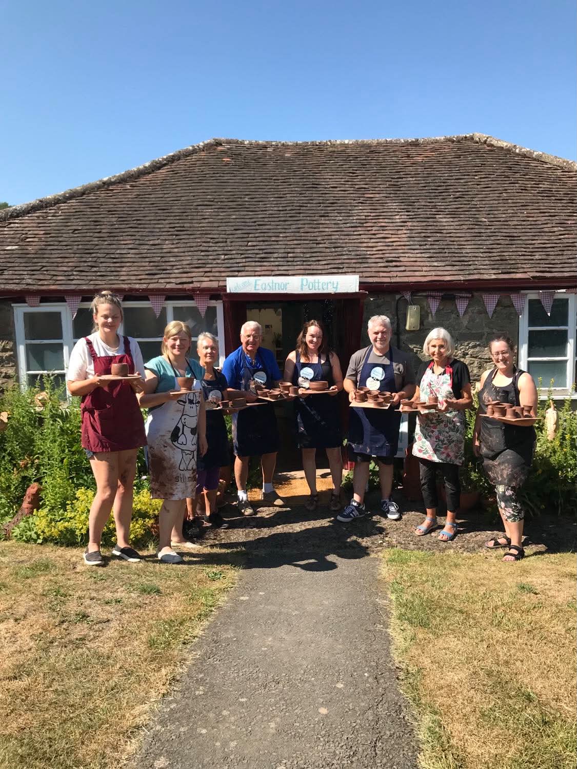 Eight adults, male and female in a line outside eastnor pottery, each person holding a board of wet brown pots that had been made on the potter's wheel. The adults are participants on a one day potter's wheel course and they are displaying their pots at the end of the day. It's a hot day, there is blue sky above the Pottery roof and the participants are wearing t-shirts and shorts under their aprons.