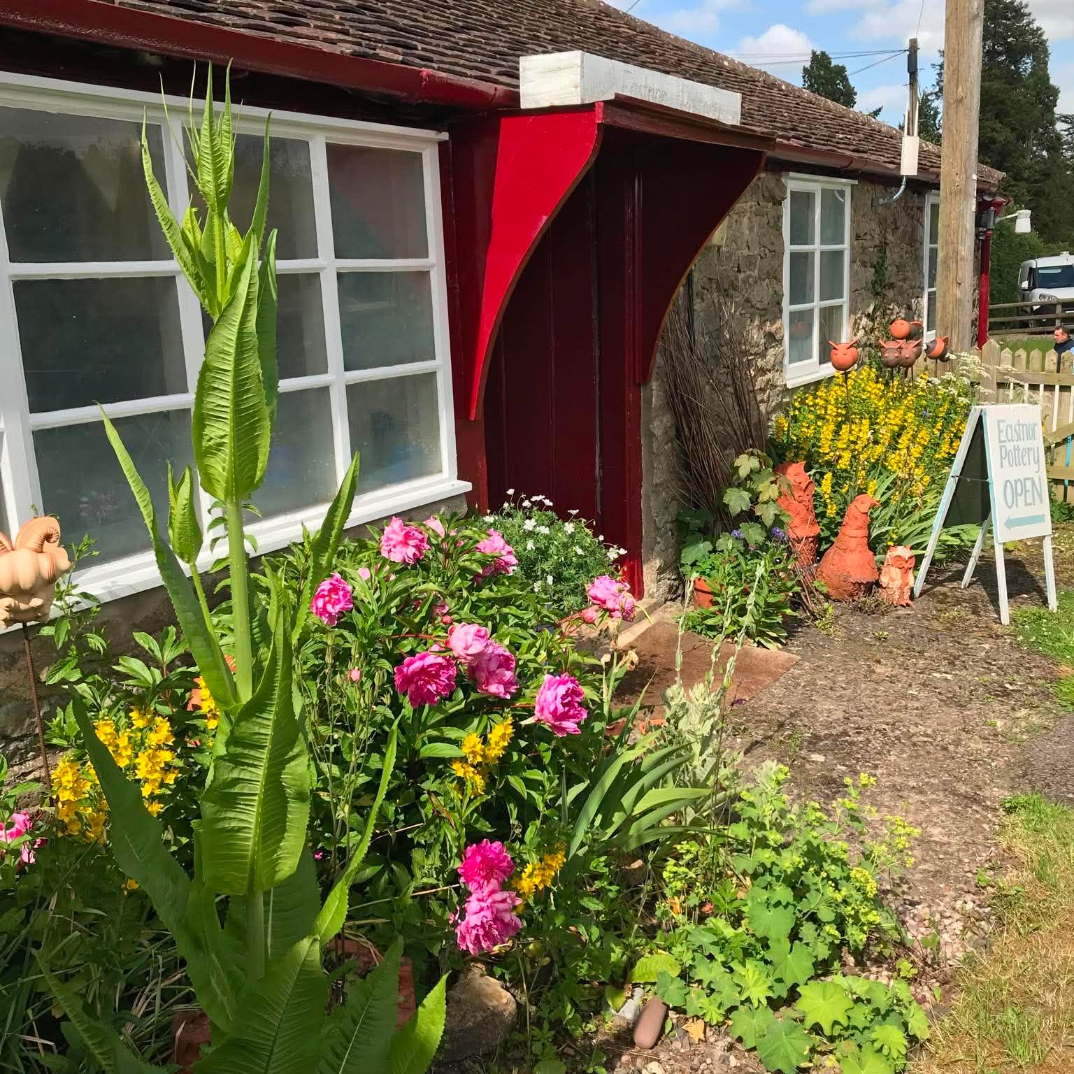Front of eastnor pottery. There is a red door and windows either side. There are pink peony flowers in the flower borders and tall yellow flowers, all very colourful. There are also some insect and bee sculptures in the flower borders outside the window made by jon williams