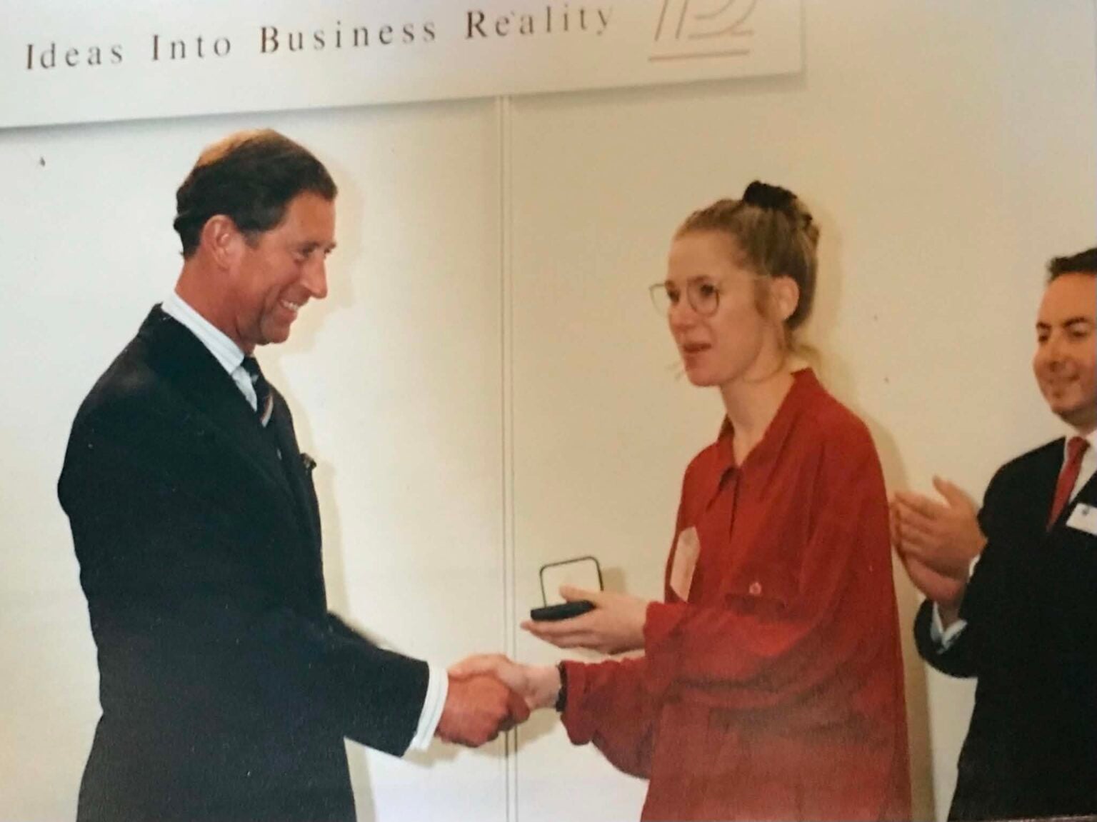 Torso and head of two figures stood in profile and shaking hands. The figure on the left is HRH king Charles and the figure on the right is Sarah Monk in a red shirt. Sarah is holding her gold medal in her left hand having just been presented to her by the king or the prince of Wales as he was known back in 1994