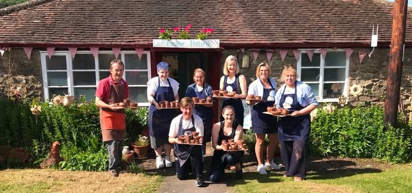 8 people wearing aprons, stood outside Eastnor Pottery's front door. They are all holding a board of pots and look very happy.
