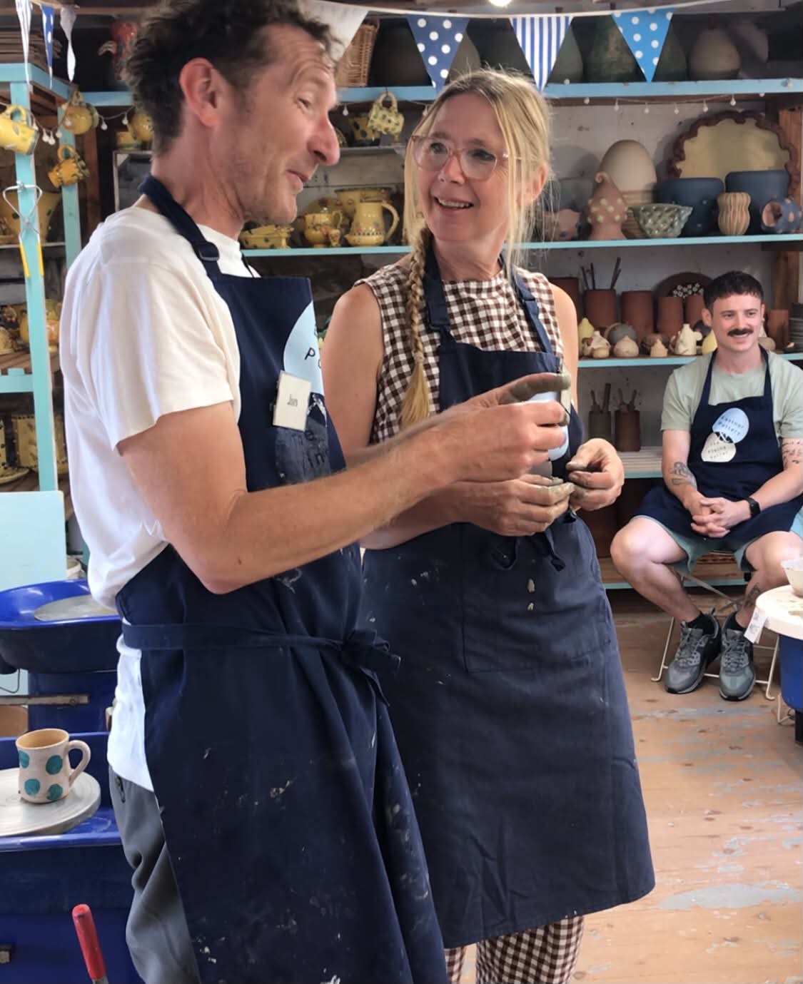 Jon williams and Sarah Monk from Eastnor Pottery discussing the work of one of their Potter's Wheel course participants. Sarah is looking at Jon who is looking down at the participant. The pair are animated in discussion and Sarah is smiling.