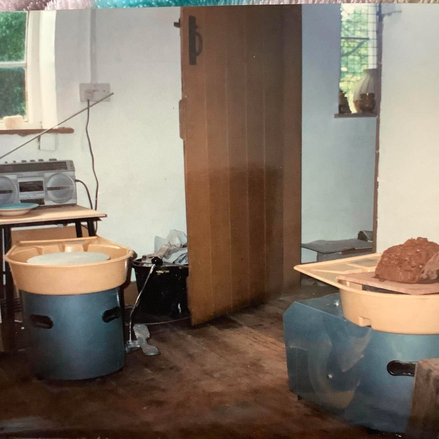 Two shimpo potter's wheels at eastnor pottery. The wheels are positioned either side of an old brown interior door. There is also a silver stereo cassette player on a table infront of the potter's wheel on the left