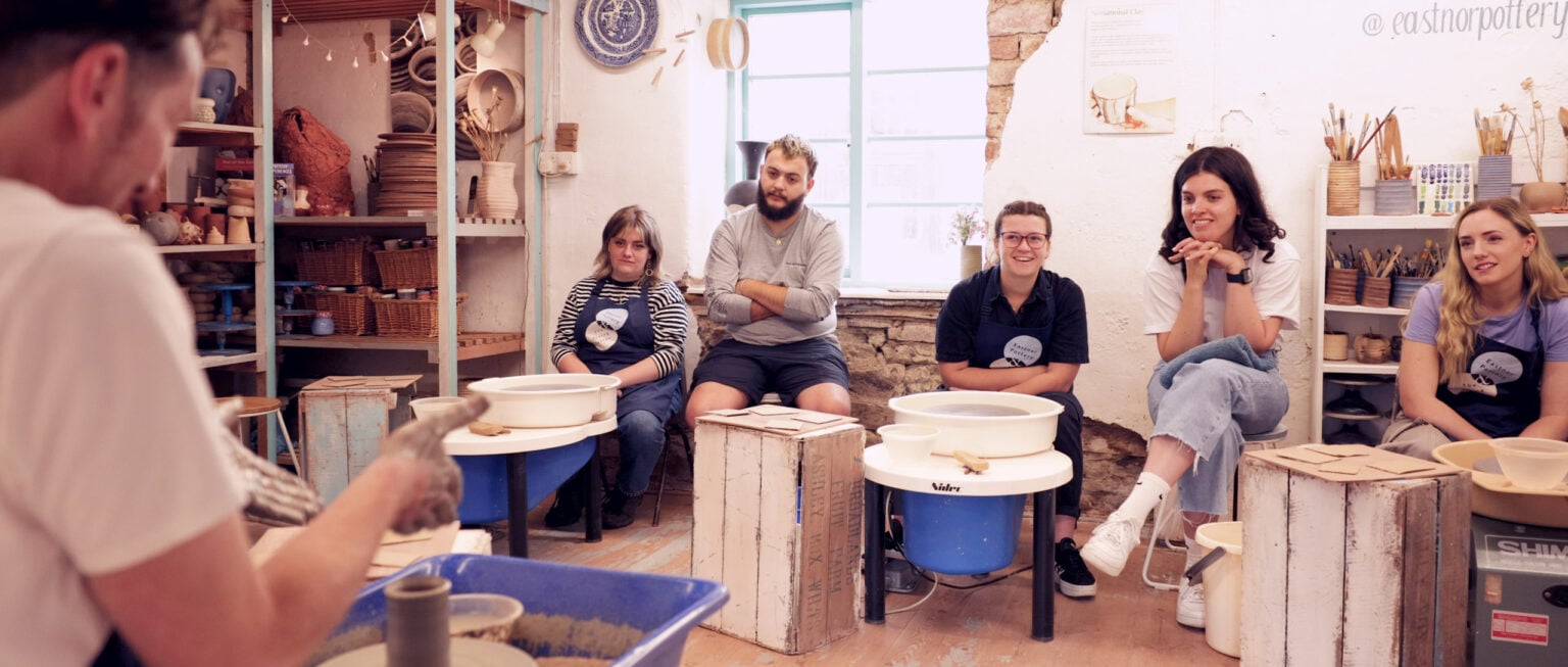 The back shoulder of the pottery tutor sat at the wheel making a cylinder pot, whilst others are looking towards him as he shows them how to make the pot.