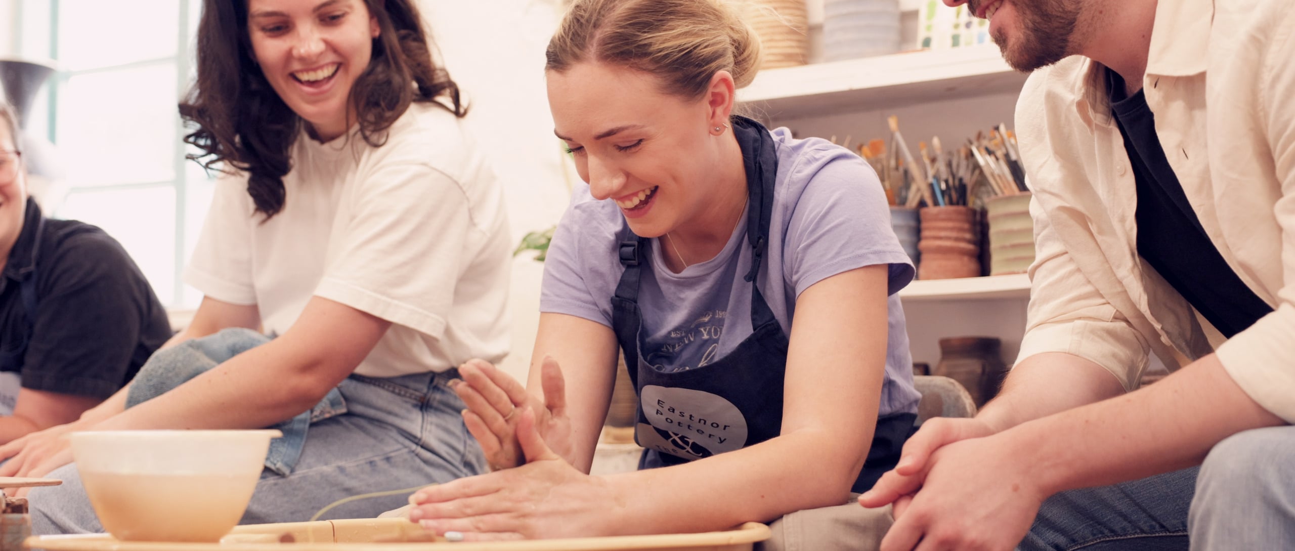 A young lady sat at the potter's wheel with others sar next to her, looking on, smiling.
