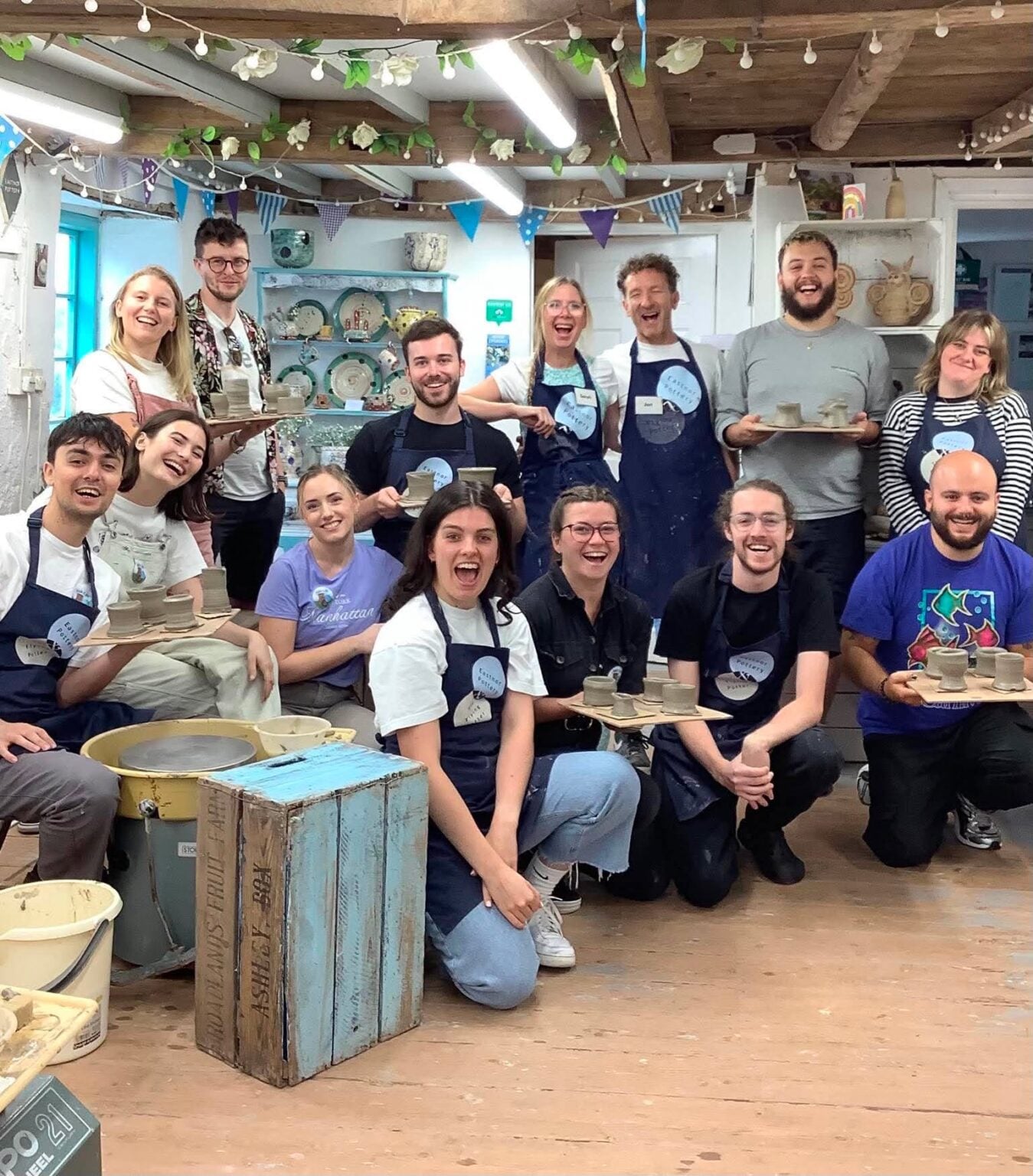 14 happy people standing in a group some are stood, some sat, some kneeling. They are wearing Eastnor pottery aprons , some are holding boards of freshly thrown pottery.