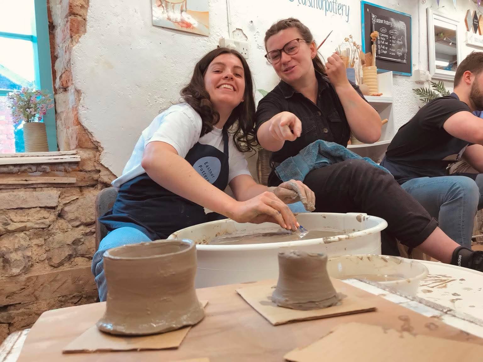 Two young women sat by a pottery wheel. One is pointing at the two pots in front of them that they have just made. They are laughing together.