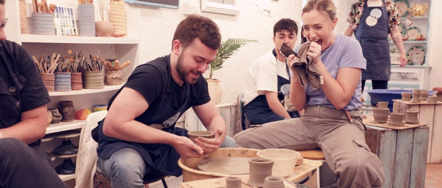 A young man is taking his pot off of the wheel whilst his girlfriend looks on with scared excitement.