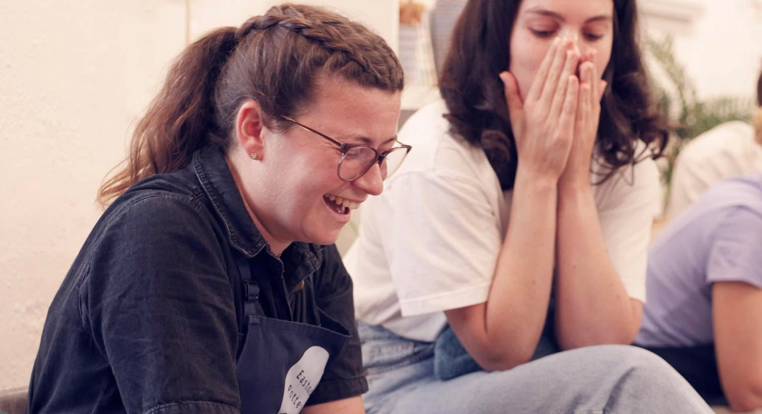 A young lady looking down and laughing, her friend by her side is covering her mouth in astonishment.