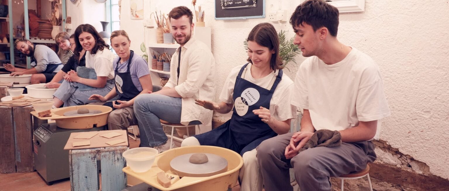A young woman placing a lump of clay on the potter's wheel others are looking on smiling.
