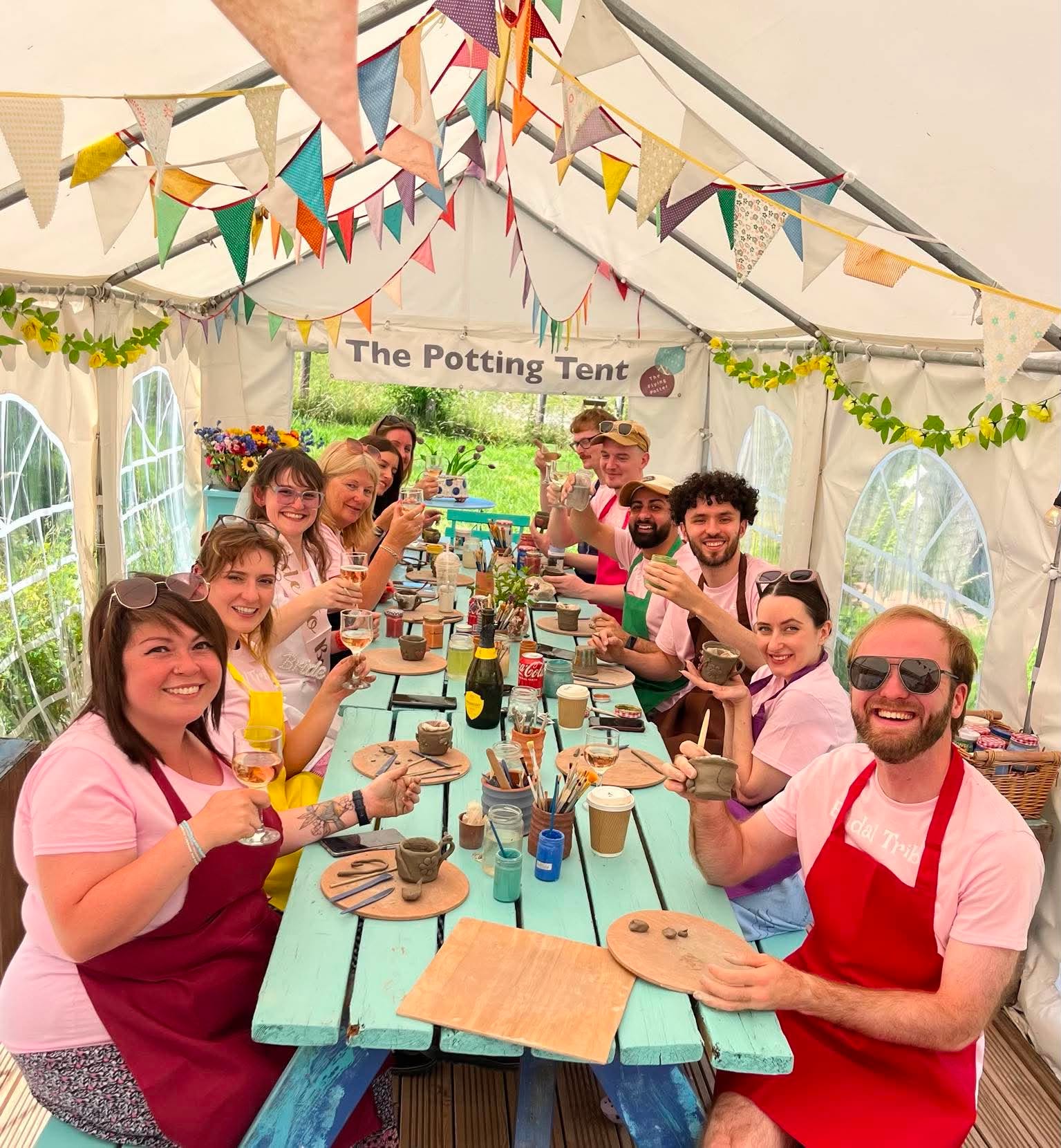 Twelve happy young people sat in a colourful marquee with a banner that says 'The Potting Tent'. They are making and decorating their own pottery . They are smiling towards the camera, some are holding up their pots, others are holding up glasses of prosecco.