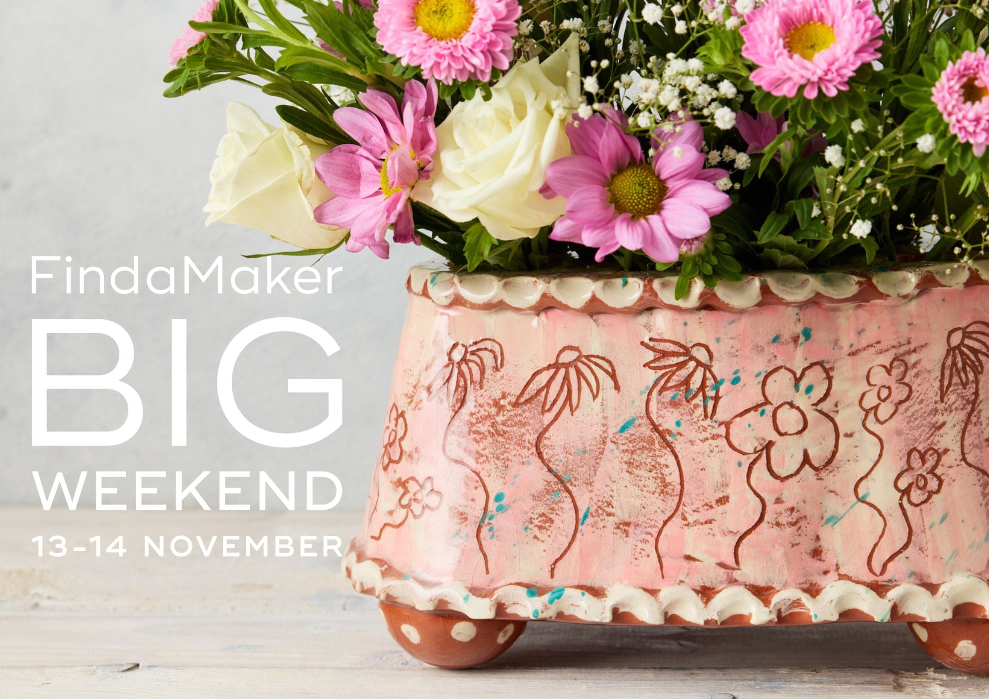 Close up of a pink pottery flower brick, oval in shape with etched sgraffito flowers as decoration. It contains fresh pink zinnias and off-white roses. There is a logo to the left that says Find a Maker Big Weekend 13 - 14 November.