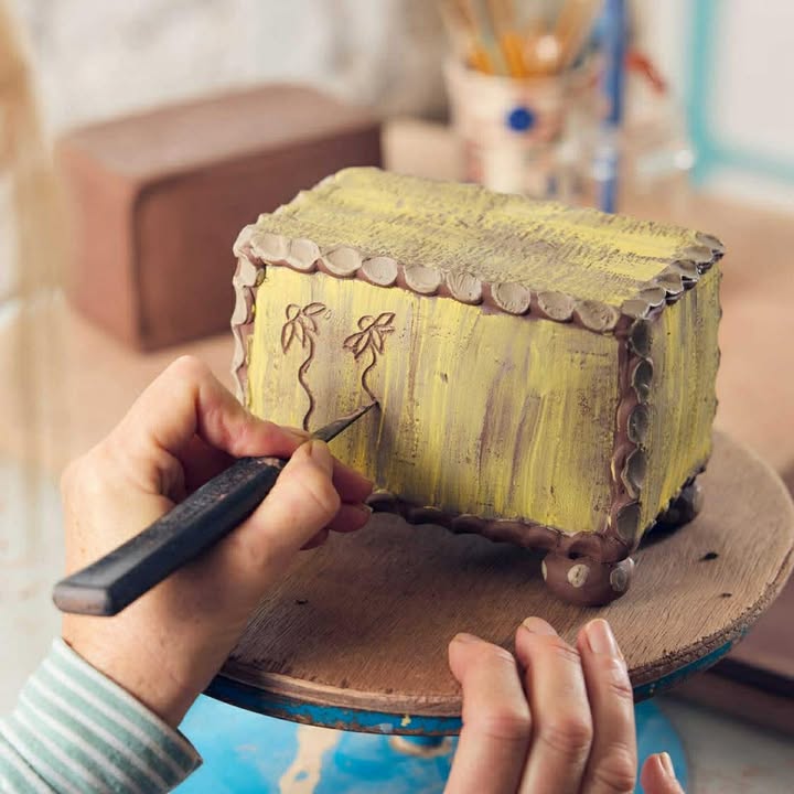 A close up photo of some hands at work, holding a pottery knife whilst using it to etch some flowers into a clay rectangular box. The box has been brushed with yellow paint too.