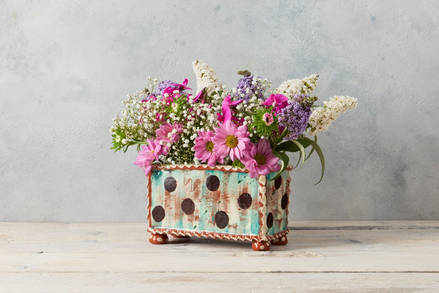 A rectangular flower brick in teal boldly decorated with large dark brown dots. It has a white piecrust edging. There is a fresh arrangement of colourful flowers including chrysanthemum, gypsophillia, and budleia.
