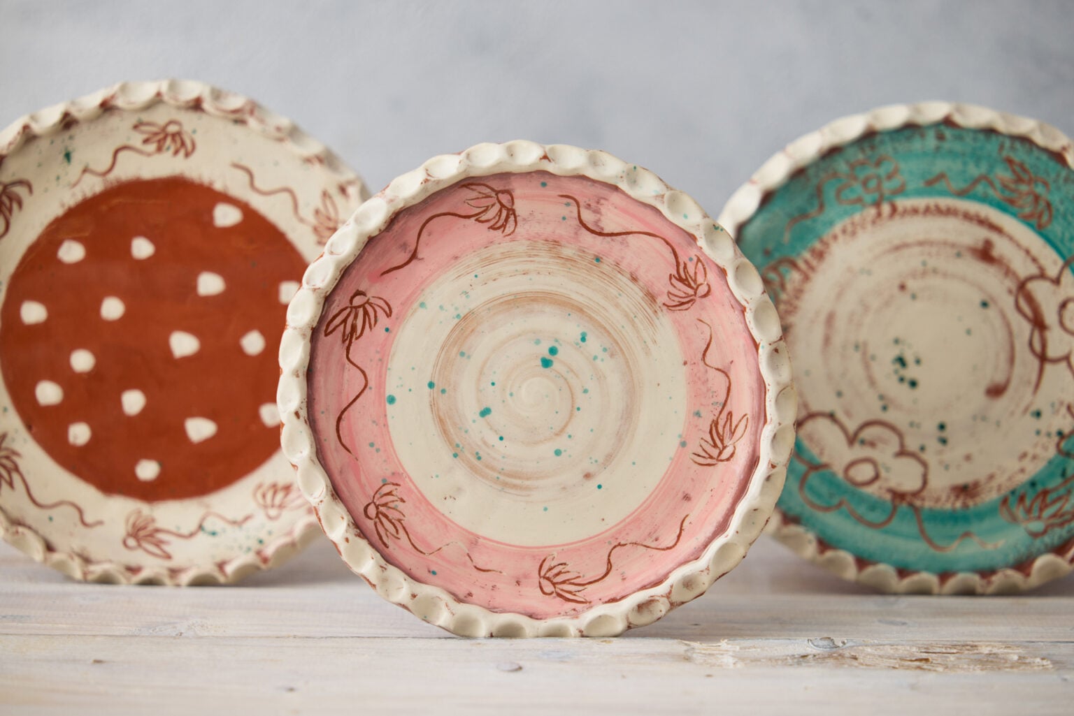 Three plates displayed upright to show the varying designs. The plate to the left is cream around the edge with etched sgraffito daisy flowers, the middle of the plate is brown with white dots. The centre plate is pink around the edge with etched sgraffito daisy flowers and is brushed cream in the centre. The plate to the right is teal around the edge with etched sgraffito large bold flowers. All three plate have a pie crust rim.
