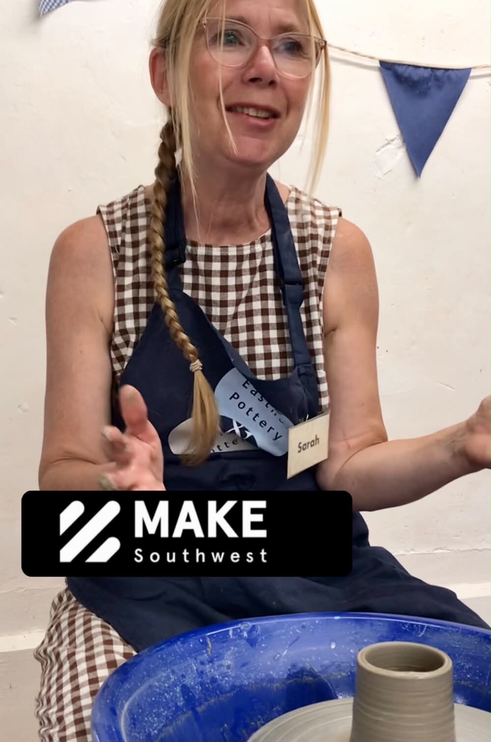 A potter at at a blue pottery wheel. She is making a pot from grey clay, it is a cylinder. She is wearing a blue apron with a name label that says Sarah. She has blond hair put into a side plait. She's looking up and her hands are up in a describing gesture. She is teaching.