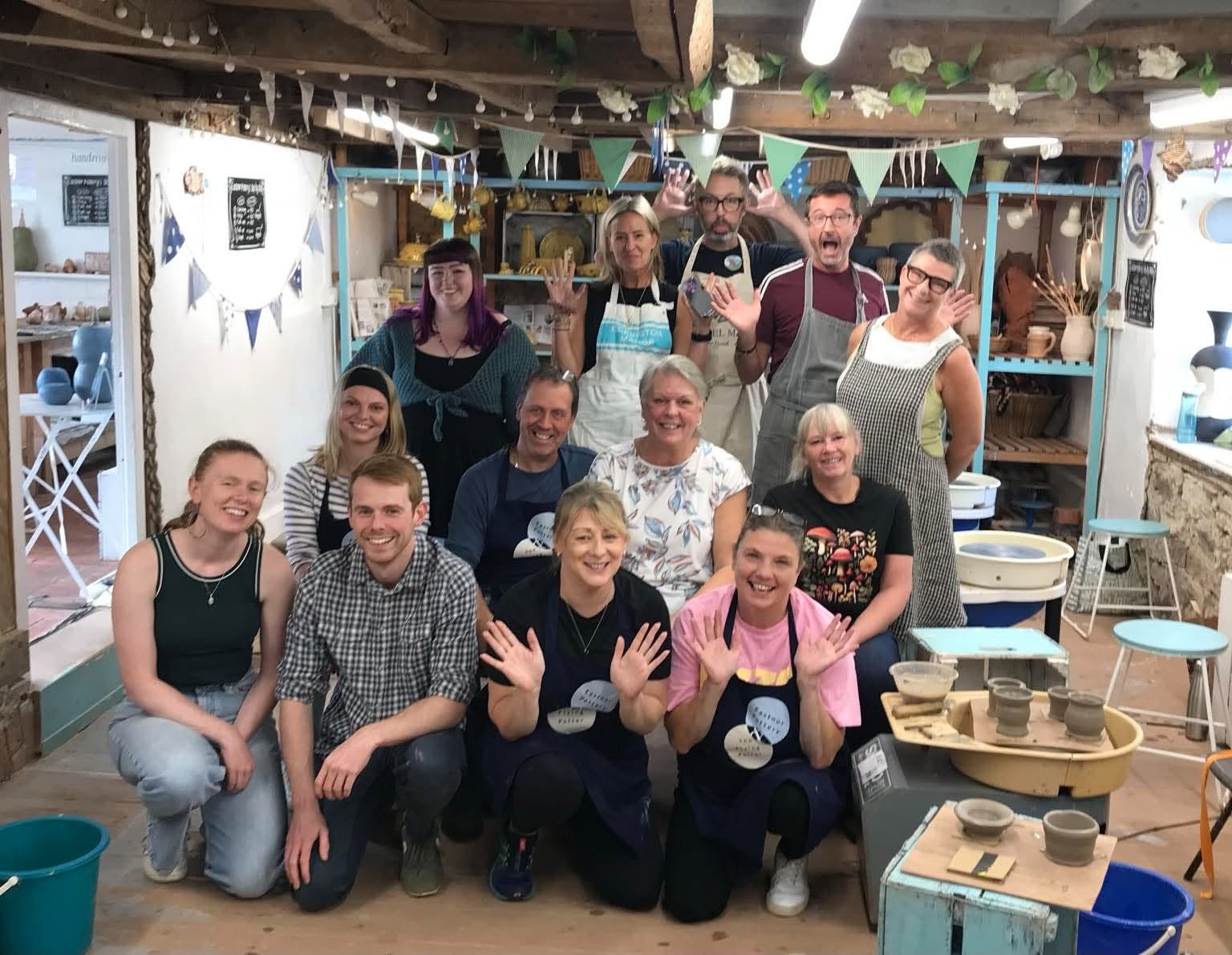 A team photo of 12 adult participants, in the studio next to the pottery wheels. A mix of men and women, a broad range of ages from 30's to 70's. Three rows of 4 people, standing, sitting and kneeling. Everybody looks very happy, some are doing 'jazz hands'!