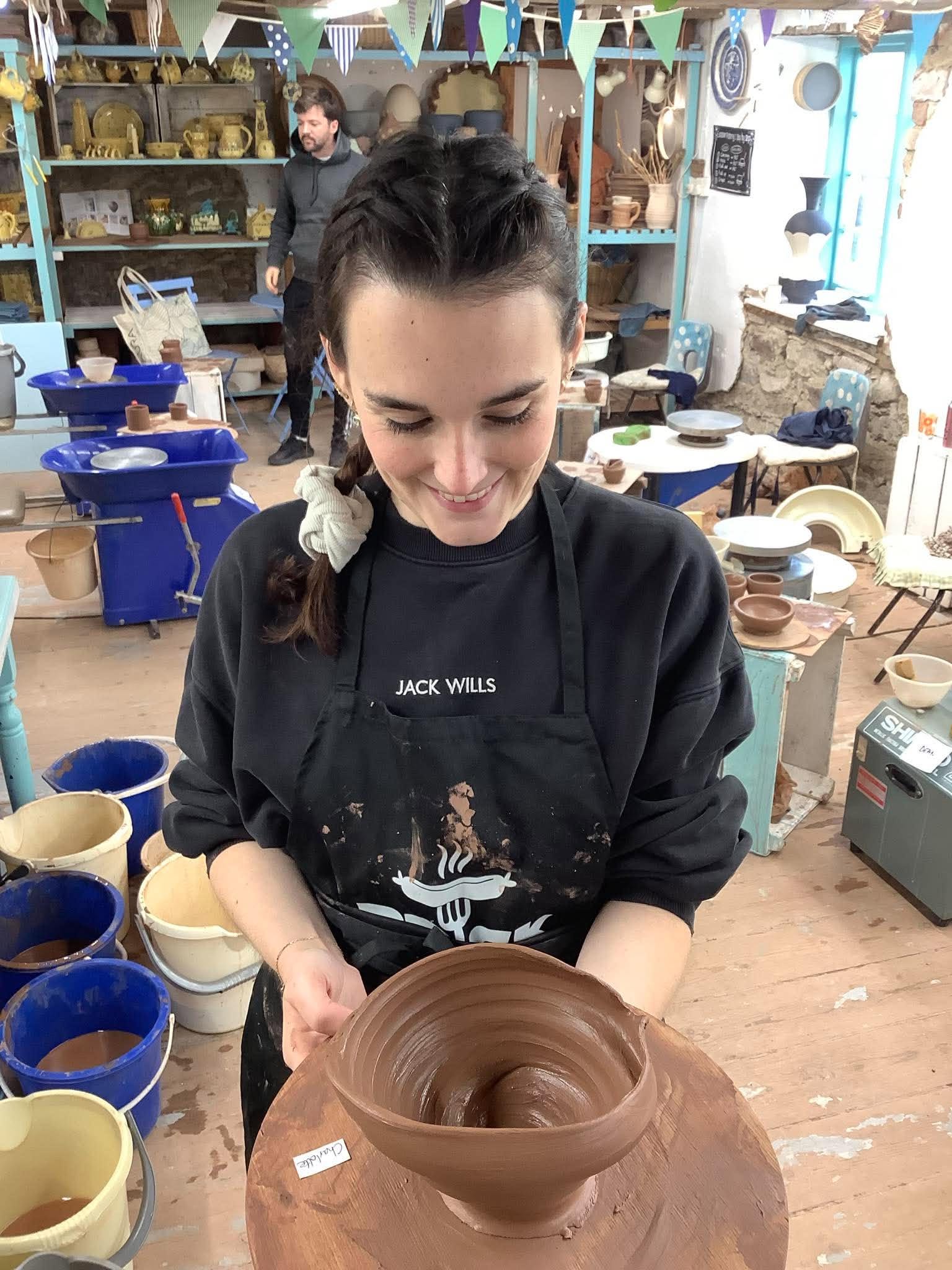 Head and shoulders of young woman holding a freshly thrown wet brown bowl thrown on the potter's wheel. She is wearing a black top, smiling and looking down at her pot on the board